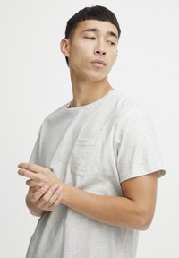 Tricou din bumbac gri deschis, cu guler rotund și un buzunar pe piept. Are pete albastre și negre împrăștiate. Mâneci scurte, croială lejeră.