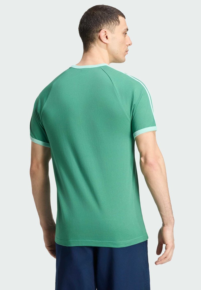 Homme aux cheveux courts et foncés portant une chemise verte à manches courtes avec une bordure bleu clair et un pantalon bleu marine, vu de dos sur un fond uni.