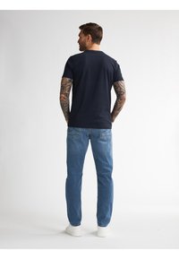 Granatowa koszulka z krótkim rękawem i okrągłym dekoltem w połączeniu z jasnoniebieskimi dżinsami, stojąca na białym tle. Widoczne białe trampki.