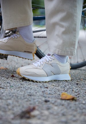 Person trägt beige und weiße Sneakers mit weißen Schnürsenkeln und Socken, ruht die Füße nahe eines Fahrradreifens auf einem Kiesweg im Freien.
