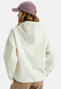 Witte fleece hoodie met een capuchon, met een gestructureerde afwerking. Gepaarde met een lichtpaarse corduroy pet. Achteraanzicht met een relaxte pasvorm.