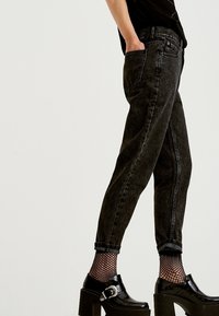 Person som bär upprullade svarta tvättade jeans, fiskenätsstrumpor och svarta plattformsskor med silverfäste, stående med ena handen i fickan.