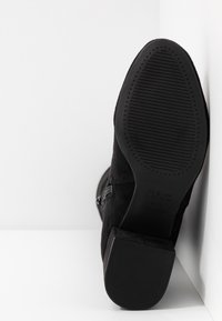 Botín de ante negro hasta el tobillo con un tacón grueso, con una suela de goma texturizada y un cierre de cremallera lateral. Logo grabado en la suela.