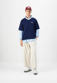 Jeune homme portant une casquette rouge, une chemise à rayures bleues avec un gilet en tricot bleu marine, un pantalon large crème et des baskets blanches, se tenant devant un fond blanc uni.