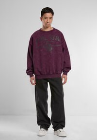 Felpa oversized viola con stampa grafica, che presenta il testo "KARL KANI" e un disegno di aquila. Abbinata a pantaloni neri oversize e scarpe bianche.