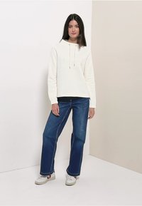 Weißer Kapuzenpullover mit Kordelzug und Logodetail, kombiniert mit dunkelblauen Jeans mit weitem Bein. Weiße Turnschuhe runden das Outfit ab.