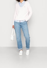 Pull en tricot rose clair avec un col en V, superposé à une chemise rayée bleue et blanche. Un jean bleu clair et des baskets blanches complètent le look.