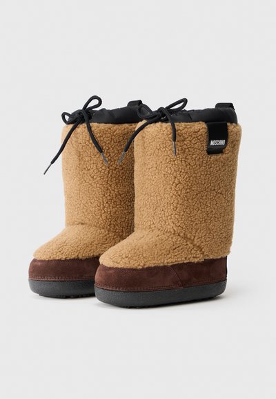 Bottes en fausse fourrure beige avec une base en suède marron, bout arrondi, partie supérieure à lacets noirs et étiquette de logo à l'arrière. Design texturé et chaleureux pour l'hiver.