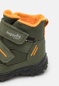 Zöld gyermekcsizma narancssárga gyapjú béléssel, tépőzárral, texturált anyagból készült, gumitalppal, amelyen tapadási minták találhatók. GORE-TEX matrica látható.