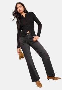 Schwarze gerippte Strickjacke mit durchgehender Knopfleiste, kombiniert mit hochgeschnittenen schwarzen Jeans und braunen spitzen Ankle Boots. Hält eine beige Handtasche.