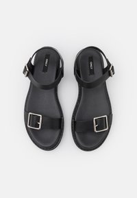 Černé kožené sandály s dvěma popruhy a čtvercovou stříbrnou sponou. Podešev je hladká a má pevnou texturovanou podrážku.