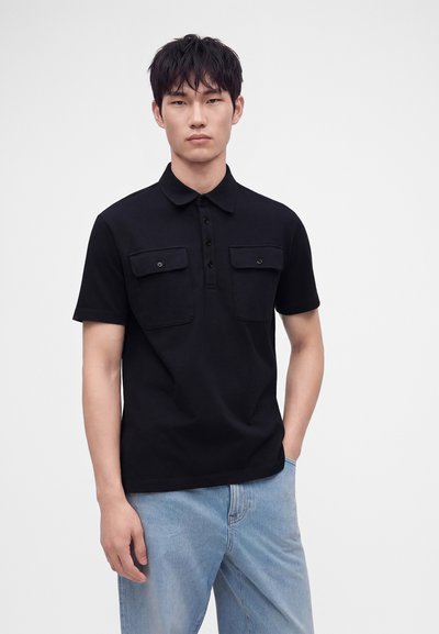 Jeune homme aux cheveux noirs courts, debout sur un fond blanc, portant un polo noir à manches courtes avec des poches avant et un jean bleu clair.