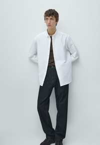 Weißes Hemd über einem braunen T-Shirt mit Rundhalsausschnitt, kombiniert mit dunklen Jeans und braunen Sneakers. Glatter Stoff, lockere Passform.