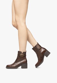 Botas de tobillo de cuero marrón con puntera redondeada, tacón cuadrado y cremallera lateral. Presentan una hebilla decorativa cerca del tobillo. Textura suave.