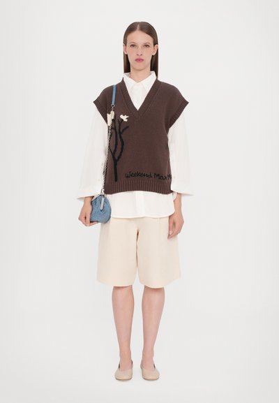 Brauner gestrickter ärmelloser Pullover mit V-Ausschnitt und floralem Design, über einem weißen Hemd und kombiniert mit beigen Shorts und einer blauen Handtasche.