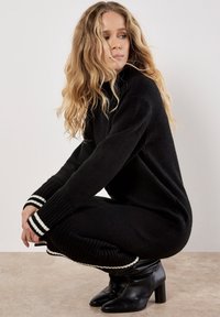 Schwarzes Strickoutfit mit hohem Kragen und gestreiften Akzenten an den Bündchen, kombiniert mit schwarzen Stiefeletten mit Absatz. Die Textur wirkt weich und gemütlich.