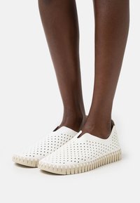 Zapatillas blancas sin cordones con diseño perforado, suela de goma beige y costura en zigzag en el borde. Textura suave, material liviano.