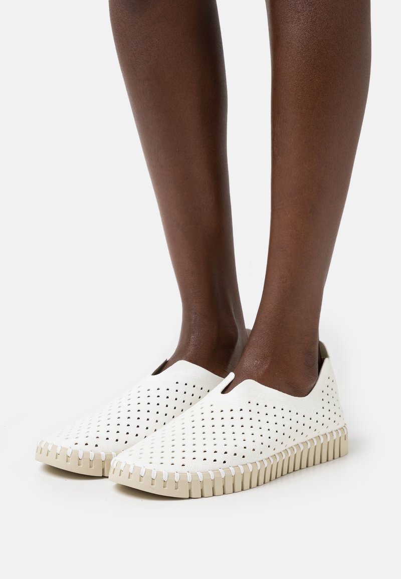 Zapatillas blancas sin cordones con diseño perforado, suela de goma beige y costura en zigzag en el borde. Textura suave, material liviano.