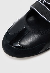 Calçado preto com acabamento em verniz liso, detalhes em camurça e uma ampla tira elástica com riscas brancas. Design com biqueira arredondada.