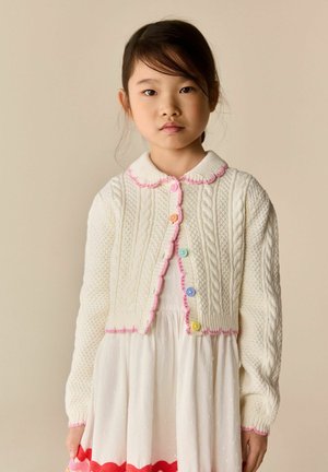 Young girl wearing a white textured cardigan with colorful buttons and pink trim over a white dress, standing against a beige background.