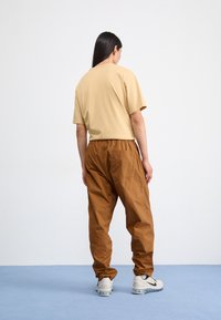 Homokszínű pamut póló barna gumis derekú nadrággal párosítva. Az öltözet laza szabású, és világos színű sportcipőkkel van kiegészítve.