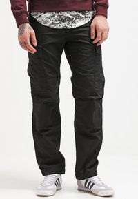 Pantalons cargo noirs avec plusieurs poches, fabriqués à partir d'un tissu résistant. Associés à des baskets blanches avec des bandes noires et une chemise imprimée en dessous.