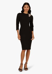 Robe noire ajustée avec des manches trois-quarts, embellissement en bijoux sur l'épaule et une longueur au genou. Tissu lisse et design moulant.