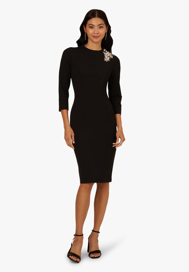 Robe noire ajustée avec des manches trois-quarts, embellissement en bijoux sur l'épaule et une longueur au genou. Tissu lisse et design moulant.