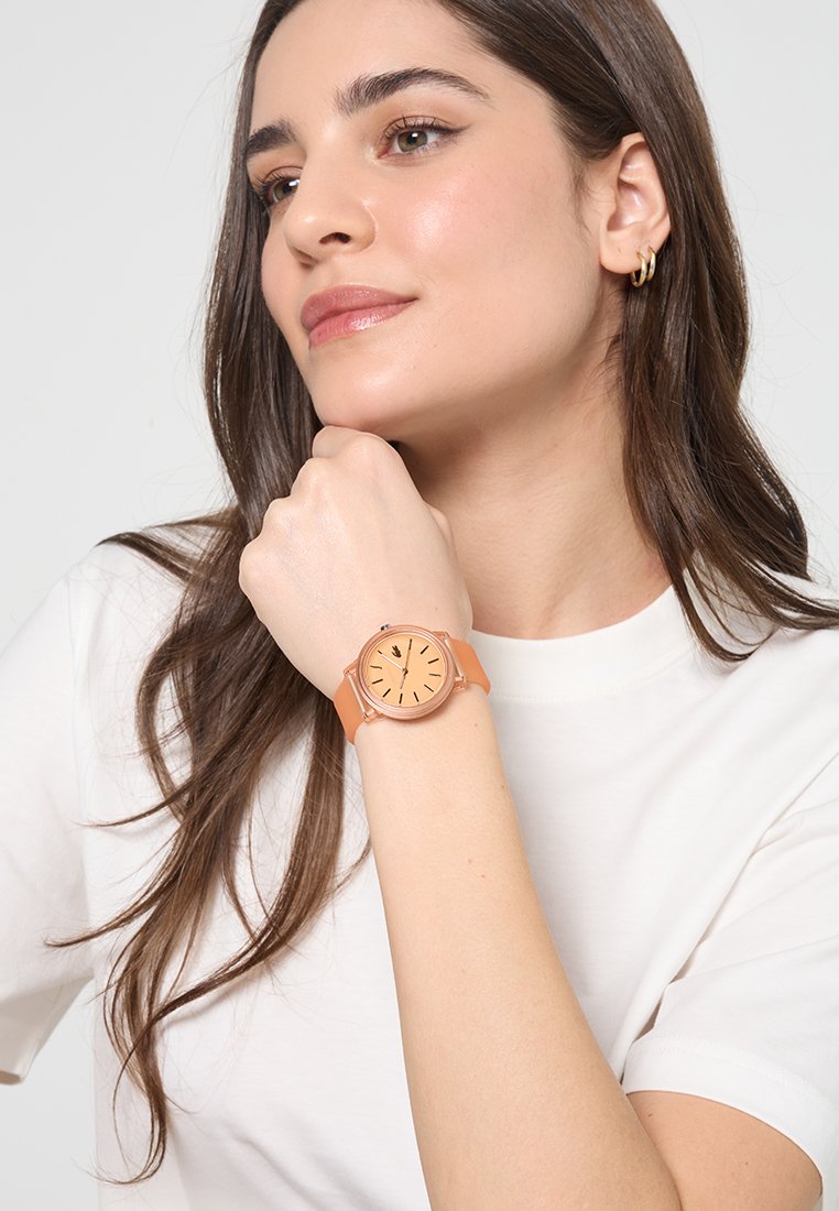 Reloj de oro rosa con cara redonda y correa de silicona naranja. Presenta marcadores y manecillas de hora negras, llevado en una muñeca sobre un fondo liso.