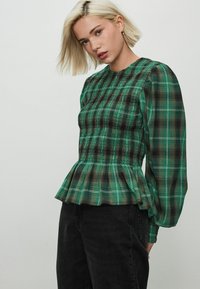 Roheline plaid blouse pikade puff-särkrigega, kogutud talje ja peplum-hemiga. Kangas on tekstuuriga ja tumedate aktsentidega.