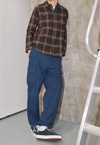 Brun rutig flanellskjorta med gröna detaljer, blå cargobyxor och flerfärgade sneakers. Står mot en betongvägg med en stege.