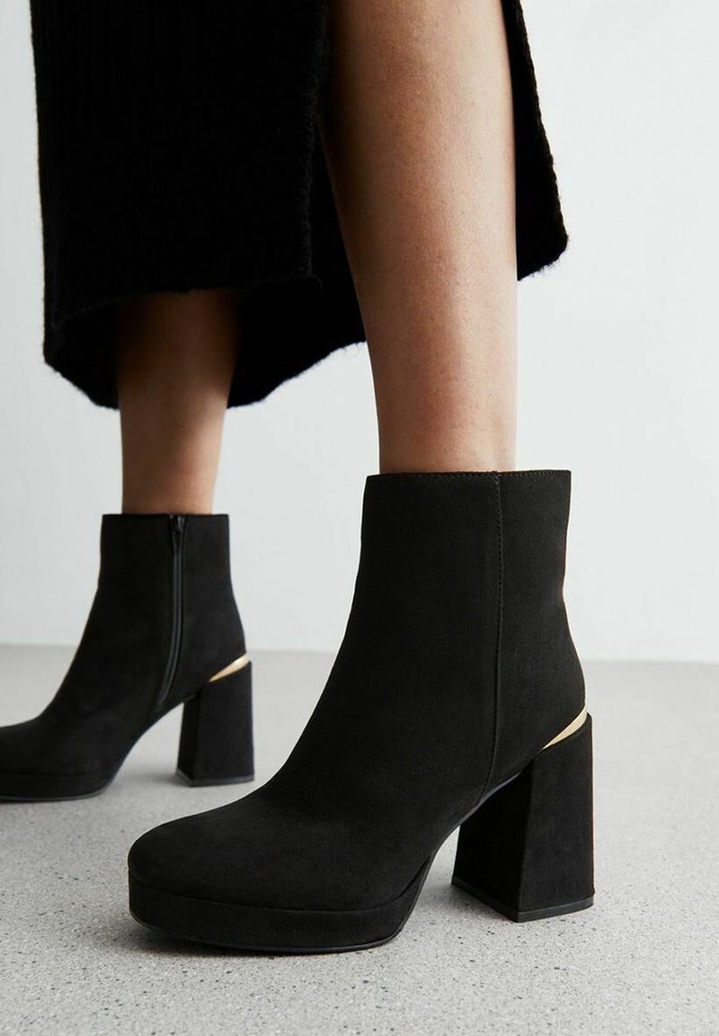 Femme portant des bottines en daim noir avec des hauts talons épais et un accent doré, se tenant sur un sol gris clair.