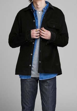 Personne portant des vêtements superposés avec une veste en velours côtelé noire sur une chemise en jean bleue et un t-shirt gris, en train de boutonner la veste.