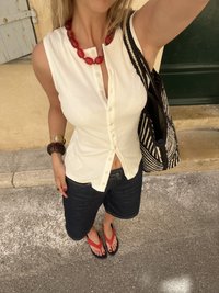 Femme portant un haut blanc sans manches boutonné, un short noir, un collier rouge, des bracelets, portant un sac rayé noir et blanc, debout à l'extérieur.