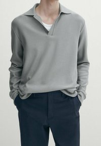 Szary sweter z dekoltem w szpic i kołnierzykiem, wykonany z teksturowanej tkaniny. W połączeniu z ciemnoniebieskimi spodniami, ręce w kieszeniach.