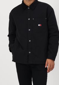 Svart knäppjacka i mjukt tyg; har en kordflordskrage, bröstficka med färgglad logotyp och silverfärgade tryckknappar.