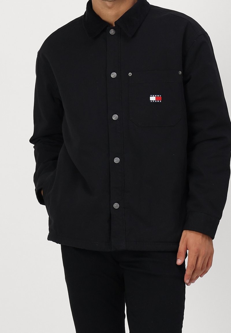 Svart knäppjacka i mjukt tyg; har en kordflordskrage, bröstficka med färgglad logotyp och silverfärgade tryckknappar.