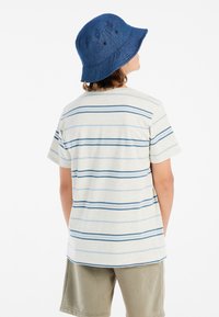 Korte mouw gestreepte T-shirt in lichtbeige met blauwe strepen, gecombineerd met gedempte groene shorts en een donkerblauwe bucket hat.