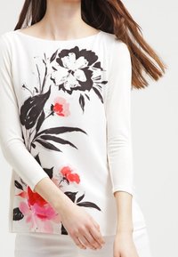 Top a maniche lunghe con stampa floreale che presenta fiori neri e rosa su sfondo bianco, con scollatura tonda e vestibilità ampia.