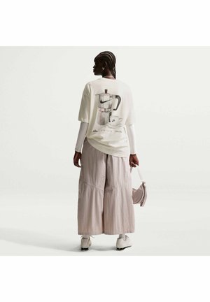 Kobieta z warkoczami, ubrana w oversize'ową białą koszulkę z nadrukiem ekspresu do kawy, szerokie beżowe spodnie, białe buty, trzymająca małą bladoróżową torebkę.