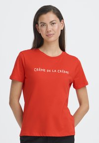 Crvena pamučna majica s natpisom "CRÉME DE LA CRÉME" u kontrastnoj boji, okrugli izrez i kratki rukavi. Glatka tekstura.