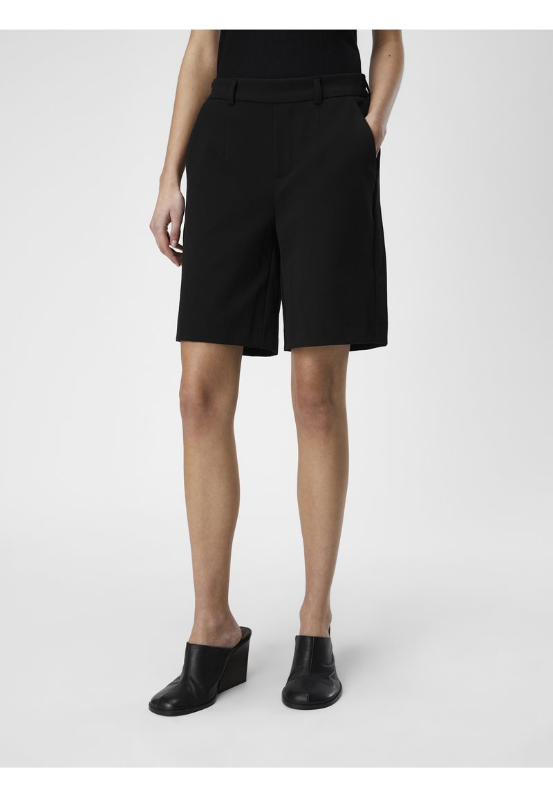 Femme portant un short noir sur mesure jusqu'aux genoux et des chaussures noires à talons compensés slip-on, debout sur un fond uni.