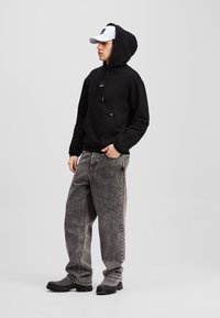 Čierna mikina s kapucňou a predným vreckom, skombinovaná s voľnými šedými džínsami a čiernymi odolnými čižmami. Biela baseballová čiapka s logom.