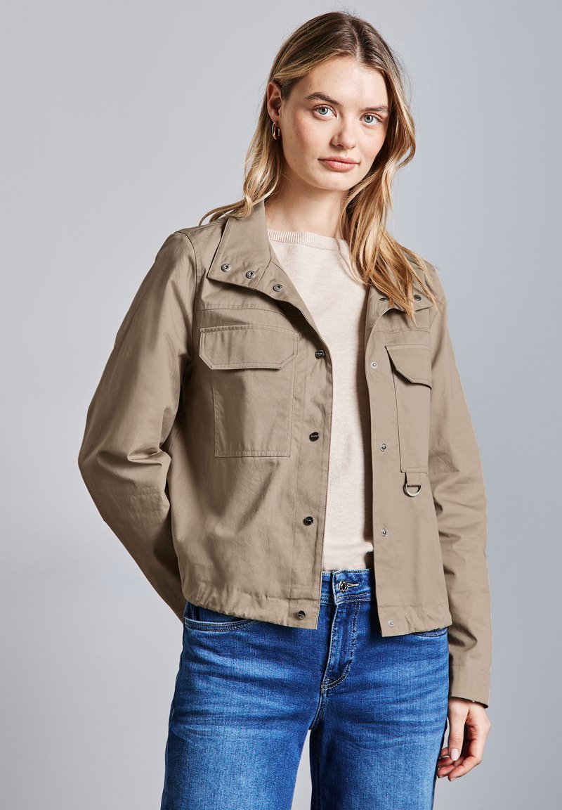 Beige kort jacka med knappstängning och fyra fickor, tillverkad av lätt material; kombinerad med en krämfärgad topp och blå jeans.