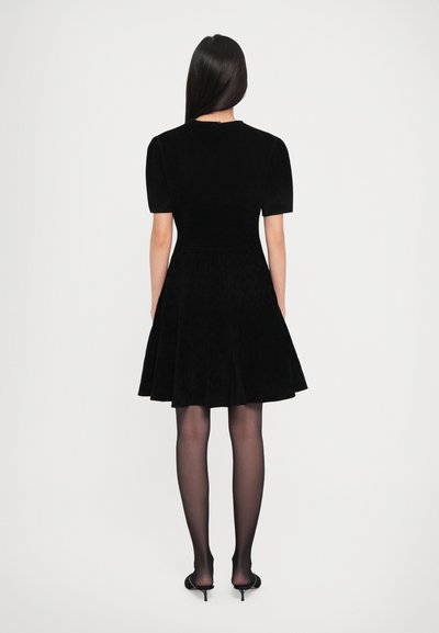 Woman facing away in short black dress with puff sleeves, sheer black tights, and black kitten heel shoes on plain white background.
