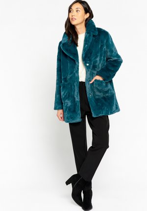 Teal faux fur coat with a large collar, two front pockets, and a relaxed fit, worn over a cream sweater and paired with black pants.