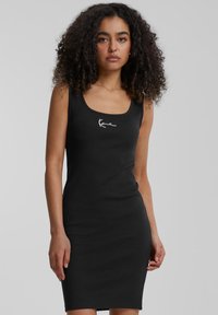 Juodas prigludęs suknelė-šalikas, pagaminta iš raibos medžiagos, su apvaliu iškirpimu. Turi baltą logotipą krūtinėje ir iki kelių siekiančią iškirpimo formą.