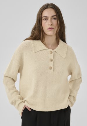 Mladá žena s dlhými hnedými vlasmi, oblečená v krémovom pletenom svetri s tromi gombíkmi a čiernych nohaviciach, stojaca na jednoduchom sivom pozadí.