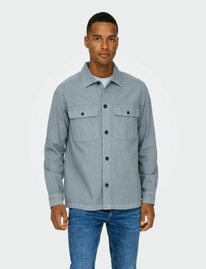 Camisa de mezclilla gris con botones en la parte delantera, dos bolsillos en el pecho y mangas largas. Usada con vaqueros azules, presenta un corte recto y un cuello clásico.
