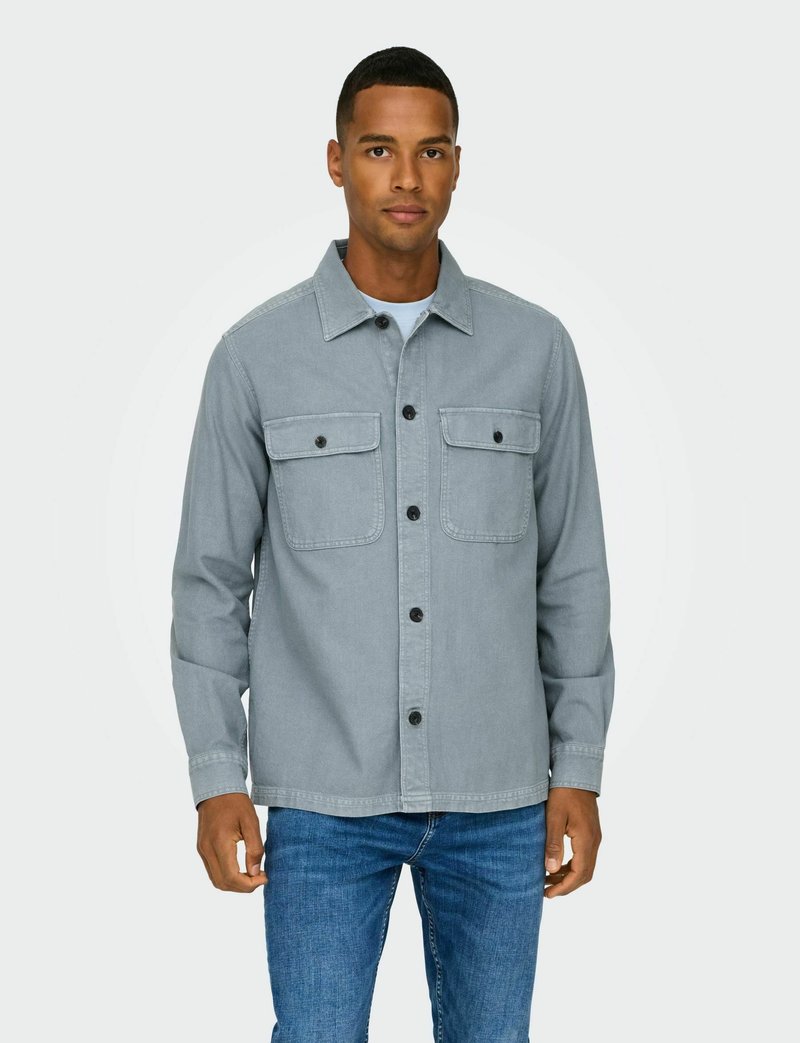 Camisa de mezclilla gris con botones en la parte delantera, dos bolsillos en el pecho y mangas largas. Usada con vaqueros azules, presenta un corte recto y un cuello clásico.